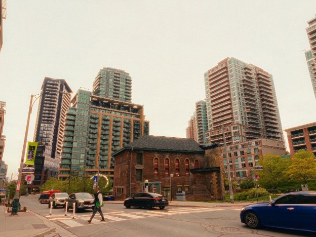 Picture8 - Toronto Bangla Town | Digital Bangla Weekly Magazine Historic brick building at a busy city intersection, flanked by modern high‑rise towers as cars pass and pedestrians cross the street.