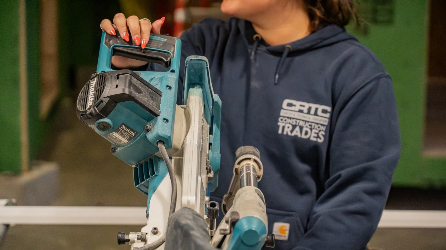 Picture8 - Toronto Bangla Town | Digital Bangla Weekly Magazine Person in a navy CRCT Construction Trades hoodie operating a teal Makita miter saw in a workshop