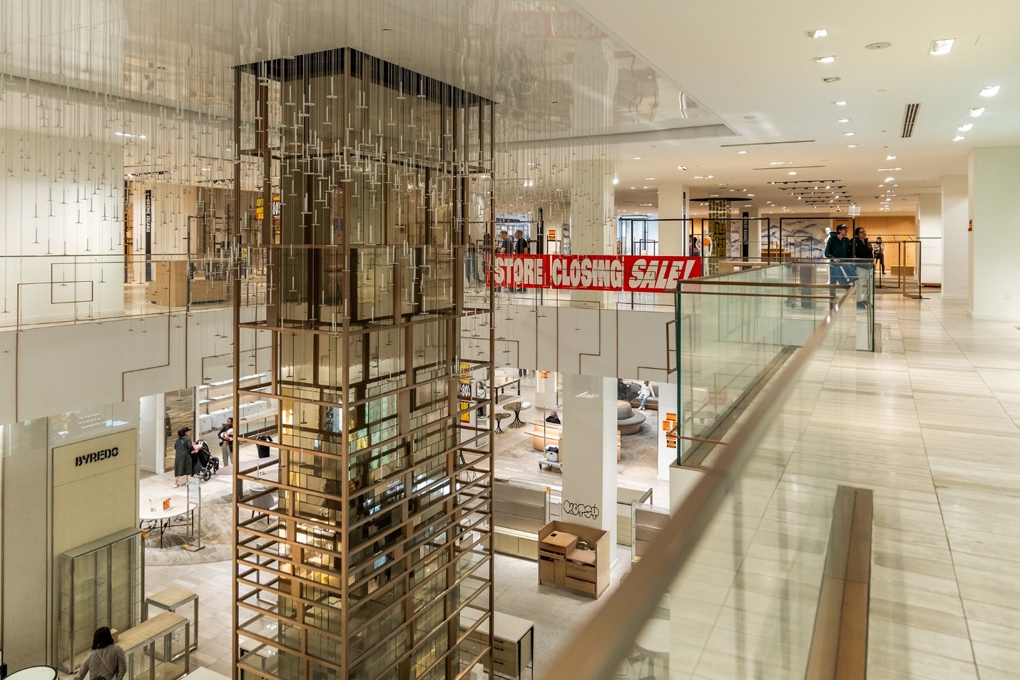 Picture9 - Toronto Bangla Town | Digital Bangla Weekly Magazine Interior of a modern shopping mall with a tall glass elevator shaft and a red 'STORE CLOSING SALE' banner across the upper level.