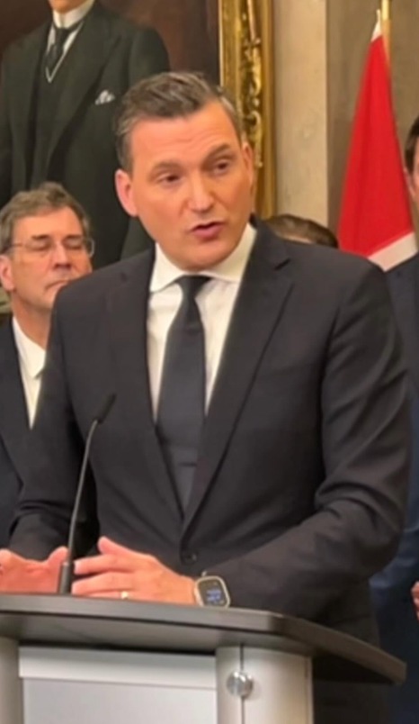 minister - Toronto Bangla Town | Digital Bangla Weekly Magazine Man in a dark suit and tie speaks at a podium with a microphone in a formal room; a red-and-white flag is visible in the background.