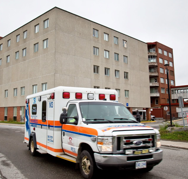7 - Toronto Bangla Town | Digital Bangla Weekly Magazine Emergency ambulance with orange and blue stripes driving past a beige, multi-story apartment building, red lights on top visible.