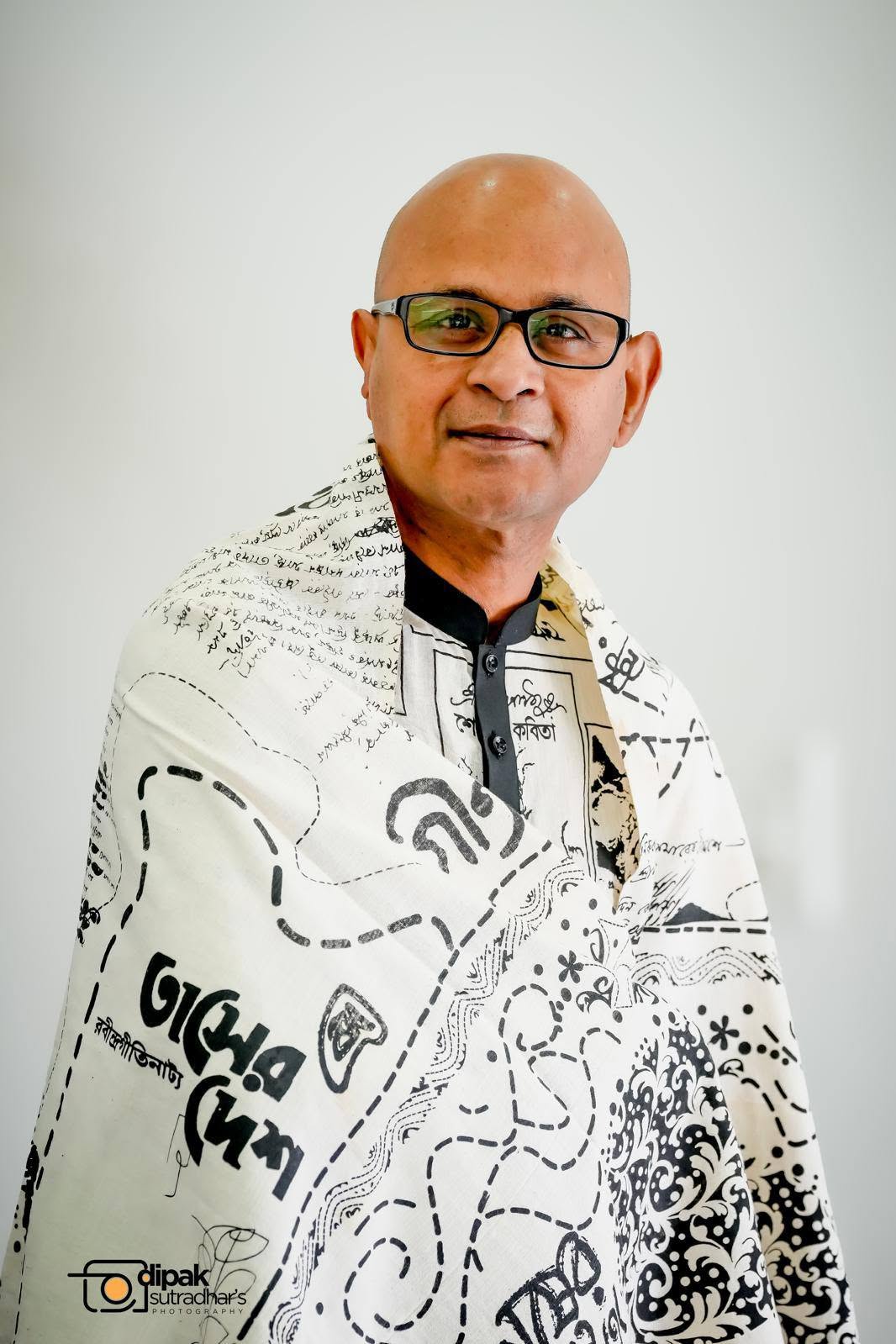 732152411_3490610317761566_4372634393468639188_njpg - Toronto Bangla Town | Digital Bangla Weekly Magazine Portrait of a bald man with glasses wearing a patterned white shirt against a light background.