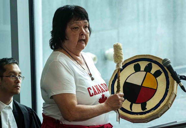 Picture1 - Toronto Bangla Town | Digital Bangla Weekly Magazine Woman in a white Canada shirt speaks into a microphone while holding a decorated hand drum.