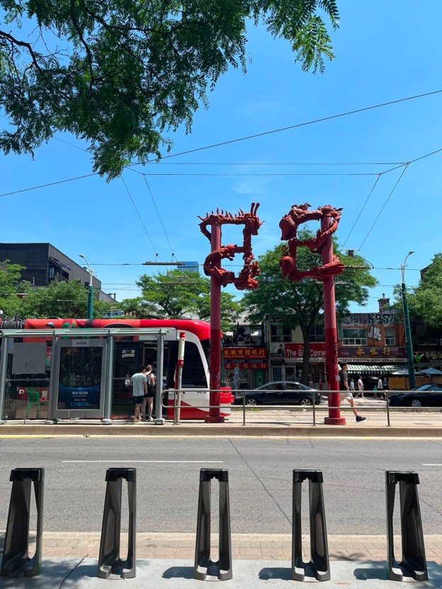 Picture1 - Toronto Bangla Town | Digital Bangla Weekly Magazine Two red dragon statues on tall columns frame a city tram stop with a red-and-white tram, pedestrians, and storefronts on a sunny day.