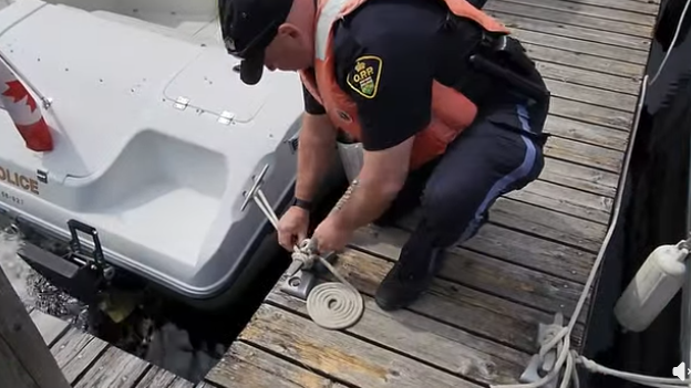 Picture1 - Toronto Bangla Town | Digital Bangla Weekly Magazine Police officer in a life vest kneeling on a wooden dock, tying or checking a mooring line beside a white rescue boat with a Canadian flag decal on the stern.