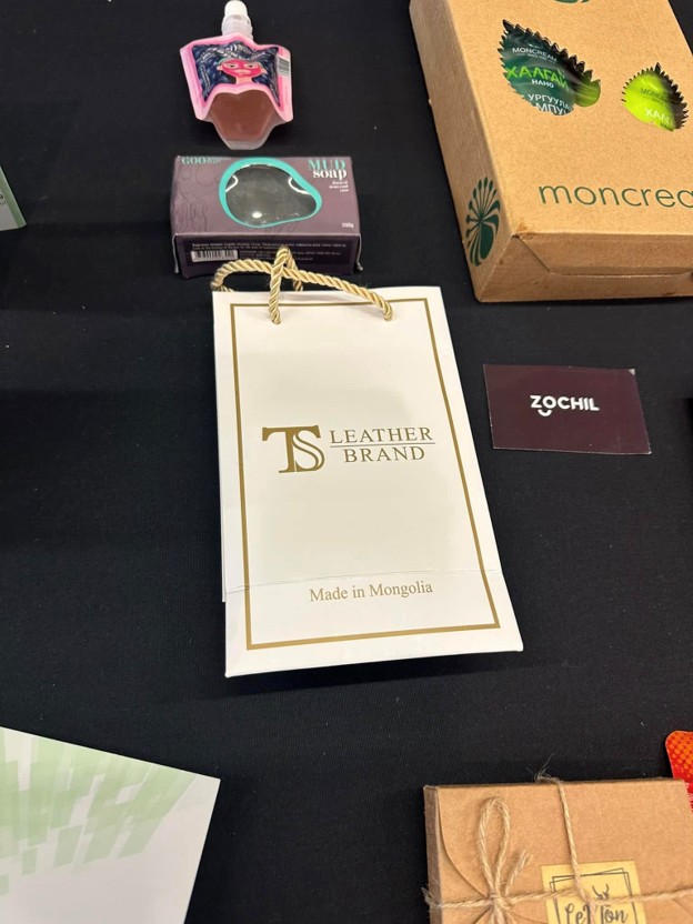 Picture10 - Toronto Bangla Town | Digital Bangla Weekly Magazine White gift bag labeled TS Leather Brand with 'Made in Mongolia' and rope handles, surrounded by small boxed products on a black surface.
