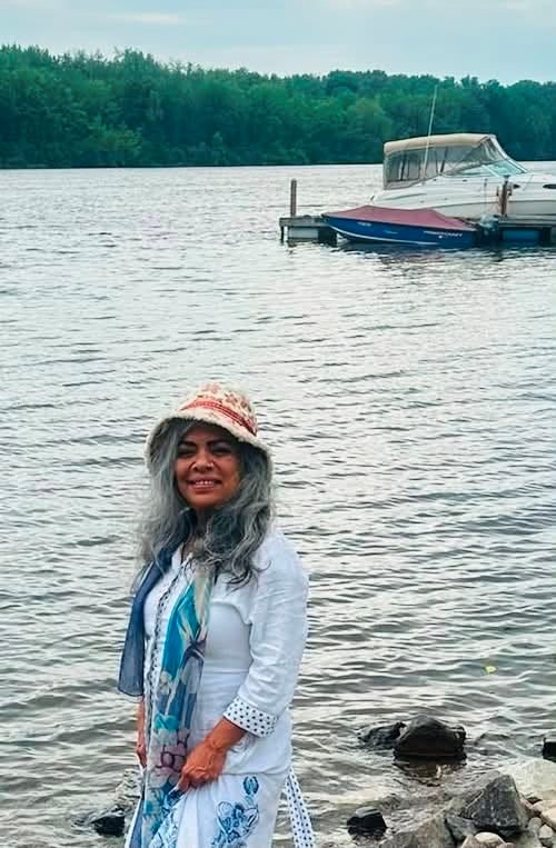 Picture11 - Toronto Bangla Town | Digital Bangla Weekly Magazine Woman with long gray hair and a straw hat standing on rocky shore by a lake, with a docked boat in the background.