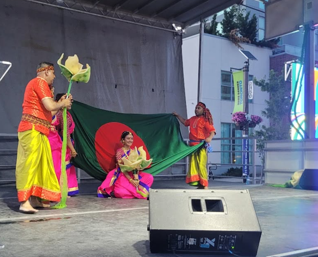 Picture12 - Toronto Bangla Town | Digital Bangla Weekly Magazine Dance troupe on an outdoor stage unrolling a large Bangladesh flag while wearing bright pink, yellow, and orange costumes with flower props central to the performance.