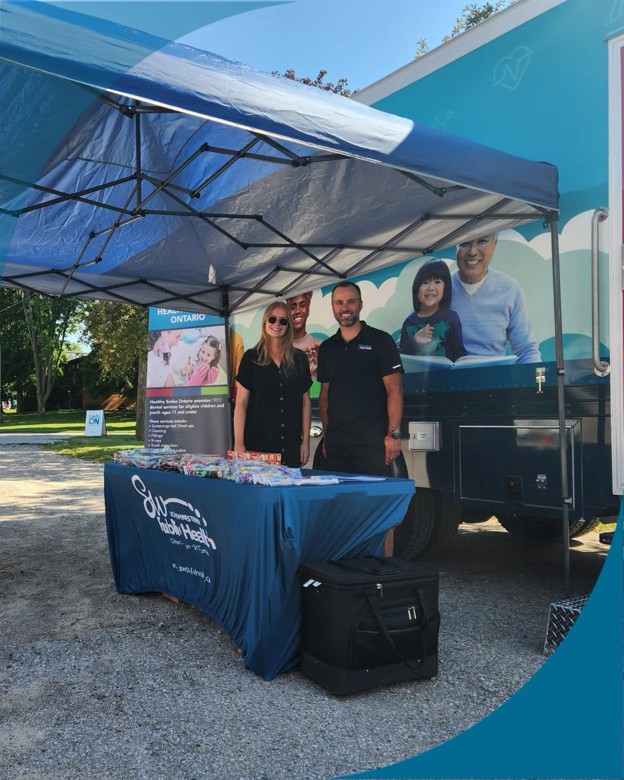 Picture2 - Toronto Bangla Town | Digital Bangla Weekly Magazine Group of three staff members at a health outreach booth under a blue canopy, with brochures on a table in front of a mobile clinic truck.