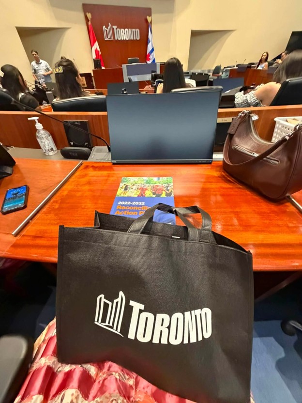 Picture2 - Toronto Bangla Town | Digital Bangla Weekly Magazine Black Toronto-branded tote bag on a wooden conference table during a meeting, with a laptop, pamphlet, and attendees in the background.