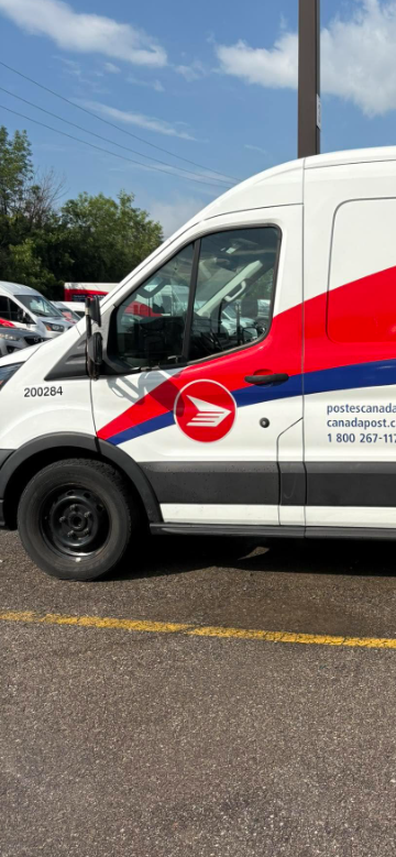 Picture2 - Toronto Bangla Town | Digital Bangla Weekly Magazine Side view of a white Canada Post delivery van with red and blue stripes and logo, parked in a parking lot.