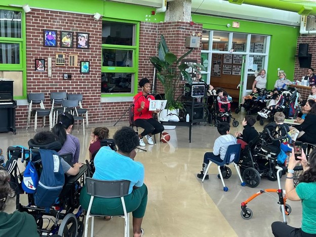 Picture3 - Toronto Bangla Town | Digital Bangla Weekly Magazine Adult in a red shirt reads a book to a diverse group of children seated in chairs and wheelchairs in a bright school room with green accents.