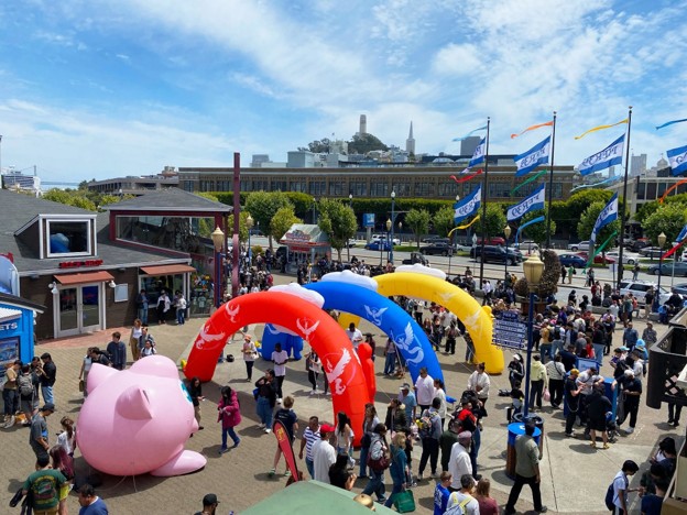 Picture3 - Toronto Bangla Town | Digital Bangla Weekly Magazine Crowd gathering at an outdoor festival with red, blue, and yellow inflatable arches and a large pink pig sculpture in the foreground.