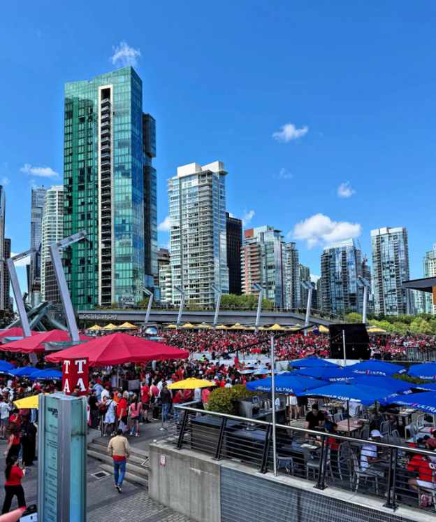 Picture3 - Toronto Bangla Town | Digital Bangla Weekly Magazine Crowd gathered at an outdoor event under blue skies, with red and blue umbrellas and glass high-rises in the background.