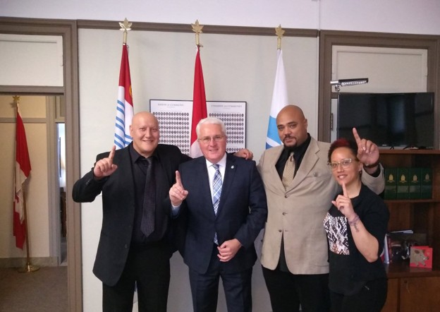 Picture5 - Toronto Bangla Town | Digital Bangla Weekly Magazine Four people in business attire stand side by side in an office, each with one index finger raised. A bald man in a dark suit, the central man in a navy suit, a bald man in a tan suit, and a woman with glasses stand in front of flags and a framed display.