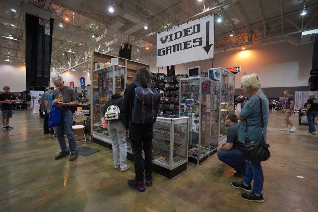 Picture6 - Toronto Bangla Town | Digital Bangla Weekly Magazine Convention floor with a 'Video Games' booth; glass display cases and shelves filled with games as attendees browse.