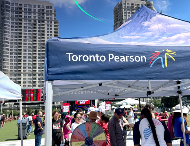 Picture6 - Toronto Bangla Town | Digital Bangla Weekly Magazine Blue Toronto Pearson tent at an outdoor event with people, a colorful wheel, and tall buildings in the background.