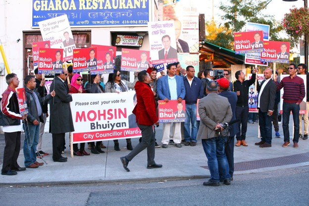 Picture7 - Toronto Bangla Town | Digital Bangla Weekly Magazine A street campaign scene with a crowd of supporters holding red-and-white signs urging votes for a candidate, outside a restaurant.