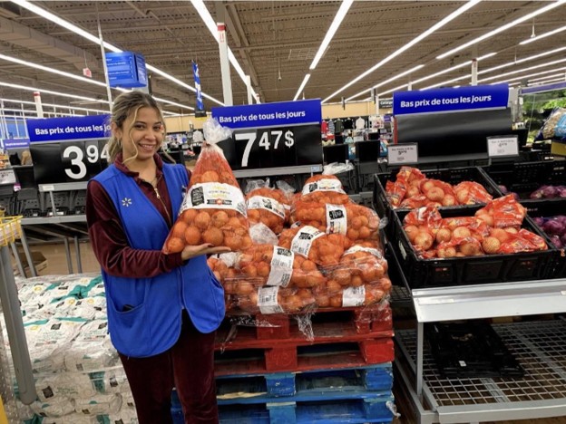 Picture8 - Toronto Bangla Town | Digital Bangla Weekly Magazine Smiling store employee in a blue vest holds a large bag of potatoes beside pallets of produce in a supermarket.