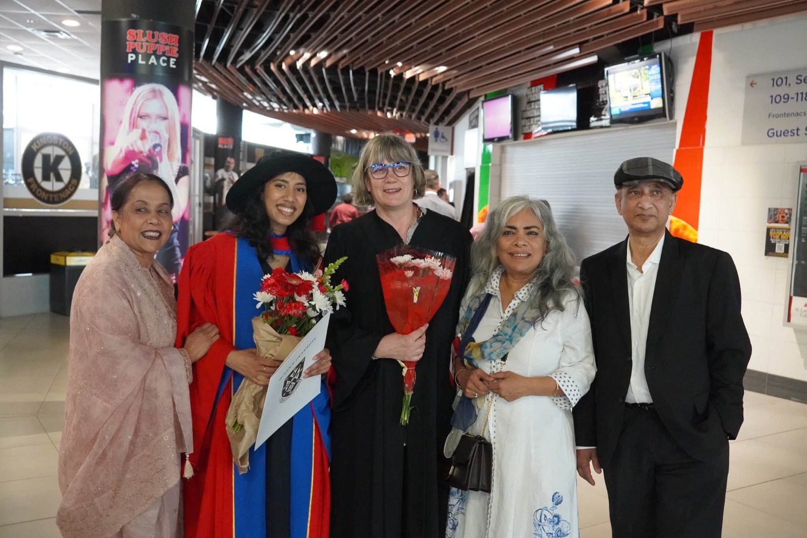 WhatsApp Image 2026-07-13 at 90300 PM - Toronto Bangla Town | Digital Bangla Weekly Magazine Five adults pose for a photo in a mall; two women in graduation gowns (red and black) hold flowers, flanked by three smiling companions in smart casual attire.